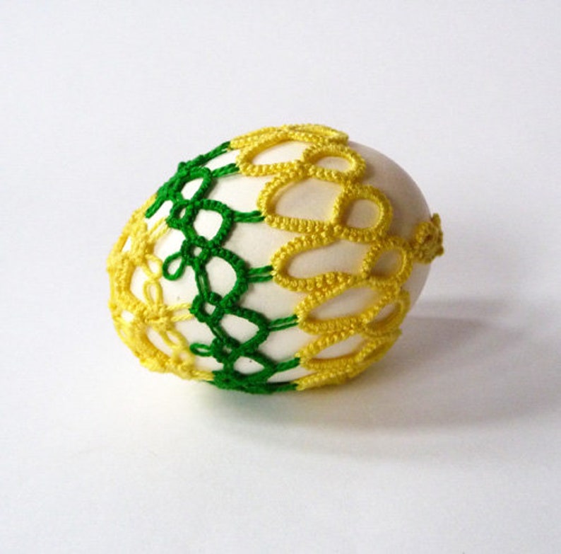 May include: A decorative egg with a white base, adorned with a crocheted overlay. The crochet design features a combination of green and yellow threads, creating a patterned, textured appearance. The egg is a craft item, likely for Easter or home decor.