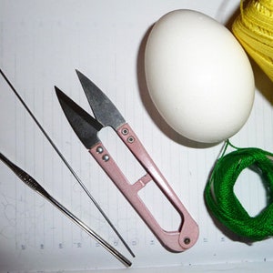 May include: A still-life image featuring crafting tools and supplies. A white egg sits next to a spool of yellow thread and a small bundle of green thread. A pair of pink-handled scissors, a crochet hook, and a needle are also present.