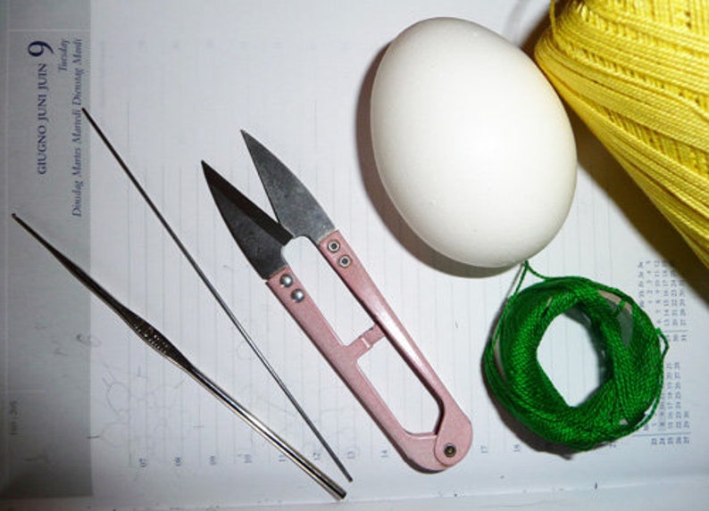 May include: A still-life image featuring crafting tools and supplies. A white egg sits next to a spool of yellow thread and a small bundle of green thread. A pair of pink-handled scissors, a crochet hook, and a needle are also present.