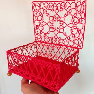 Puede incluir: Una caja de crochet roja con tapa, con intrincados diseños de encaje. La caja tiene un forro de terciopelo rojo y patas de madera.