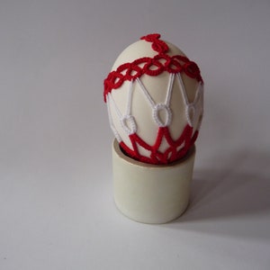 May include: An off-white egg adorned with red and white crocheted details, resting on a cylindrical stand. The egg's design features a red band around the middle and intricate white and red patterns. The stand is a light beige color.