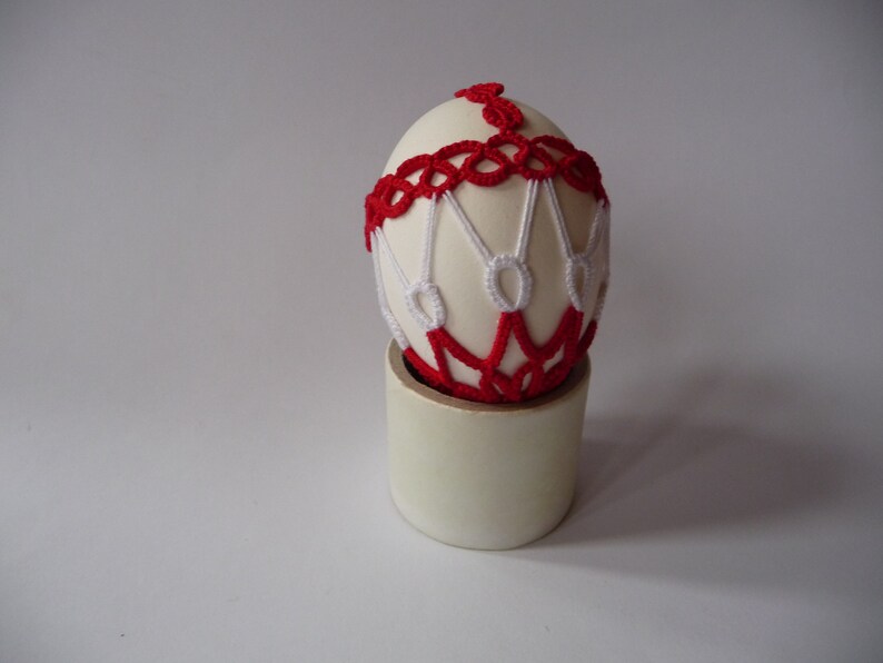 May include: An off-white egg adorned with red and white crocheted details, resting on a cylindrical stand. The egg's design features a red band around the middle and intricate white and red patterns. The stand is a light beige color.