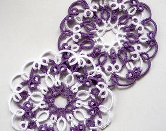 May include: Two crocheted doilies in white and purple, with a delicate floral pattern. The doilies are layered on top of each other.
