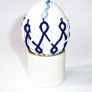 May include: A white egg-shaped decorative item with intricate blue and white woven detailing. The egg sits atop a cylindrical white base. The woven pattern encircles the egg, creating a unique visual texture. The top of the egg features a delicate blue and white woven design.