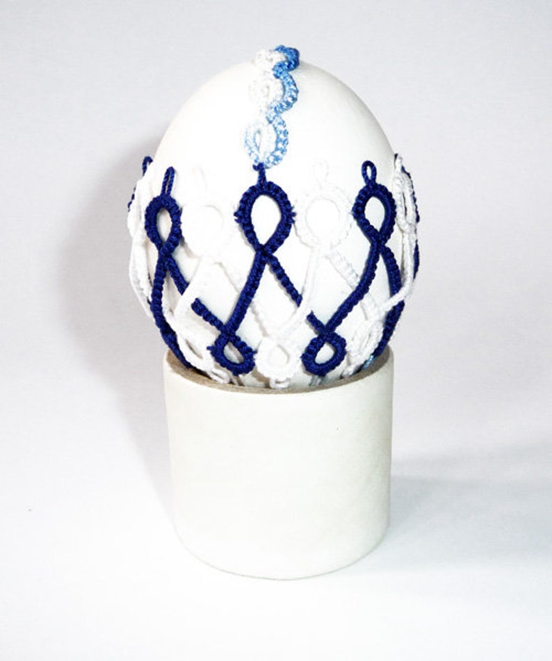 May include: A white egg-shaped decorative item with intricate blue and white woven detailing. The egg sits atop a cylindrical white base. The woven pattern encircles the egg, creating a unique visual texture. The top of the egg features a delicate blue and white woven design.