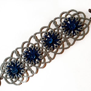 May include: A gray and blue crocheted bracelet with three blue gemstone accents. The bracelet is made with a delicate, intricate pattern.