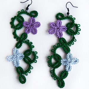 May include: A pair of green, purple, and blue crocheted earrings with floral designs. The earrings are hanging from black hooks.