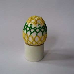 May include: A decorative egg with a white base, adorned with intricate yellow and green lace-like patterns. The egg is displayed on a small, cylindrical stand, set against a plain background. This handcrafted item is a unique decorative piece.