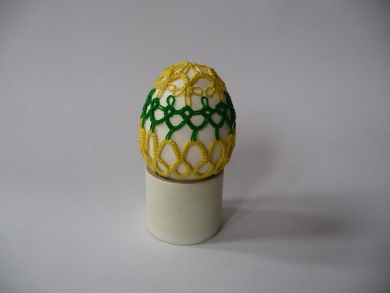 May include: A decorative egg with a white base, adorned with intricate yellow and green lace-like patterns. The egg is displayed on a small, cylindrical stand, set against a plain background. This handcrafted item is a unique decorative piece.