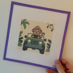 May include: A cross-stitch artwork featuring a monkey driving a green jeep. The design includes jungle foliage and musical notes on a light beige background, framed with a purple border. The artwork is held by a hand.