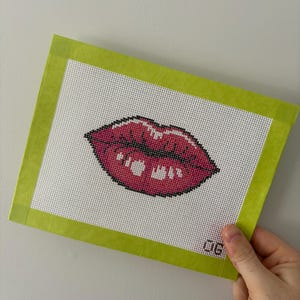 May include: A needlepoint canvas featuring a pair of red lips with a white background. The canvas is framed with a bright green border. The number "06" is visible in the bottom right corner.
