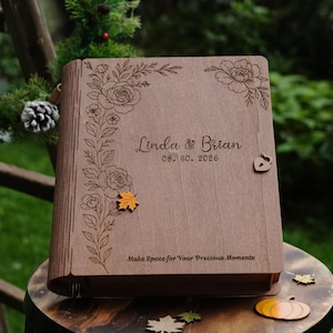 May include: A rectangular wooden photo album with a hinged spine. The cover features laser-engraved floral patterns, the names Linda & Brian, the date 08. 30. 2026, and the text Make Space for Your Precious Moments, with a small heart-shaped latch.