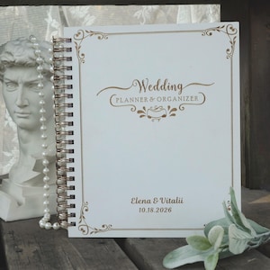 Puede incluir: Un planificador y organizador de bodas blanco con espiral, con texto dorado y borde decorativo. La portada dice "Wedding Planner & Organizer", los nombres "Elena & Vitalii" y la fecha "10.18.2026".