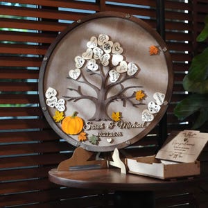 May include: A circular wooden guest book display featuring a central tree silhouette with heart-shaped wooden leaves, an orange pumpkin, and autumn-colored leaves. The text reads Sarah & Michael and 10.23.2026, with a matching wooden box nearby.