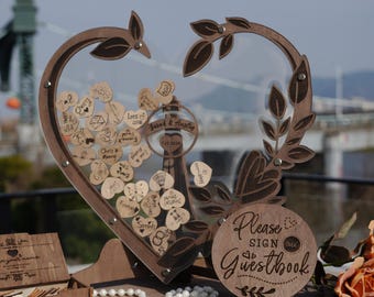 May include: A heart-shaped wooden guestbook frame with a clear acrylic front, containing small wooden hearts with handwritten messages. A central wooden circle displays the text Sarah & Austin 7.25.2025. A round wooden sign reads Please SIGN OUR Guestbook.