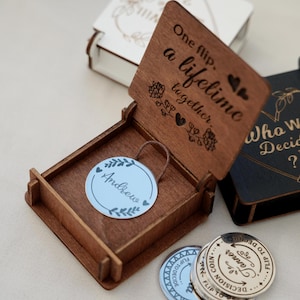 Puede incluir: Una caja de madera con tapa abierta, grabada con las palabras "One flip, a lifetime together". En el interior, una etiqueta azul claro con el nombre "Andrew" está adjunta. Varias monedas de decisión son visibles, con el texto "Flip to Decide".