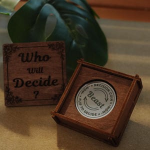 May include: A wooden decision coin set with a square box and a separate wooden square with the text "Who will Decide?". The coin is silver with the word "Beast" and "Flip to Decide".