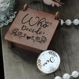 Peut inclure: Une boîte en bois avec les mots "Who will Decide?" gravés sur le couvercle, ainsi que des motifs floraux et en forme de cœur. Un petit objet rond argenté avec le nom "Jane" est à côté de la boîte. Un collier de perles est en arrière-plan.