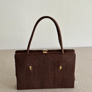 May include: A rectangular dark brown handbag with a textured, corded surface. The bag features a single arched handle, a gold-toned metal clasp closure at the top, and two small, symmetrical gold-toned wing-shaped accents on the front panel.