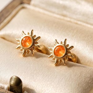 Op de afbeelding: Een paar gouden oorknopjes in de vorm van zonnen, elk met een centrale ronde oranje edelsteen. De oorbellen hebben uitstralende metalen stralen die vanuit het midden naar buiten lopen, wat een zonachtig ontwerp creëert op een crèmekleurige stof.