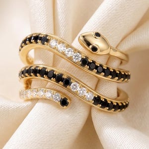 May include: A gold-toned snake-shaped ring featuring a wrap-around design. The body of the snake is encrusted with alternating clear and black stones, while the head is smooth gold-toned metal with two small black stones for the eyes.
