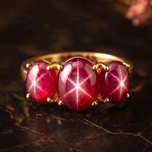 Könnte beinhalten: Ein Goldring mit drei ovalen, tiefroten Sternrubinen, die in einer Reihe angeordnet sind. Jeder Edelstein zeigt einen deutlichen sechsstrahligen Sterneffekt auf seiner glatten, im Cabochon-Schliff gearbeiteten Oberfläche. Der Ring ist aus poliertem Gold.