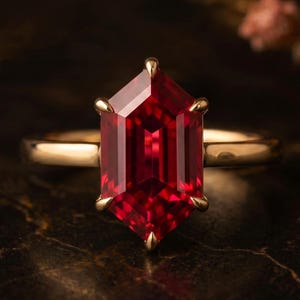 Könnte beinhalten: Ein goldener Ring mit einem großen, leuchtend roten, hexagonal geschliffenen Edelstein in einer sechs-Krappen-Fassung. Die Ringschiene ist glatt und poliert und unterstreicht mit ihrem schlichten Design die tiefroten Facetten des Steins.
