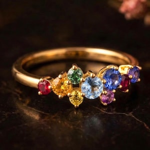 May include: A gold ring with a collection of multi-coloured gemstones. The gemstones are red, orange, yellow, green, light blue, dark blue, and purple. The ring is set against a dark background with a blurred floral element.