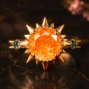 May include: A gold ring featuring a large, round, orange gemstone with a sunburst design. The ring has two smaller green gemstones on either side of the main stone. The ring is set against a dark background.