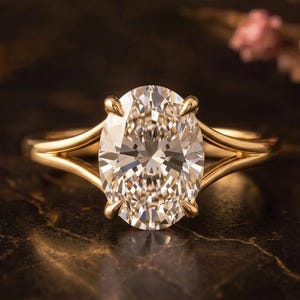 Op de afbeelding: Een gouden verlovingsring met een grote, ovaal geslepen diamant. De ring heeft een split-schacht ontwerp en vier klauwen die de edelsteen vasthouden. De diamant schittert tegen een donkere achtergrond, met zachte roze bloemen op de achtergrond.