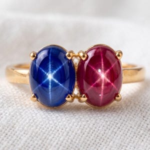 Op de afbeelding: Een gouden ring met twee naast elkaar geplaatste ovale cabochon edelstenen. De ene steen is een diepblauwe stersaffier en de andere een levendige rode sterrubijn, beide met een duidelijk zesstralig stereffect op hun gepolijste oppervlak.