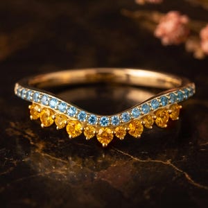 May include: A gold chevron-shaped ring featuring a row of small, round, light blue stones along the top band and a row of round, vibrant yellow stones set in a scalloped pattern along the bottom edge of the curved band design.