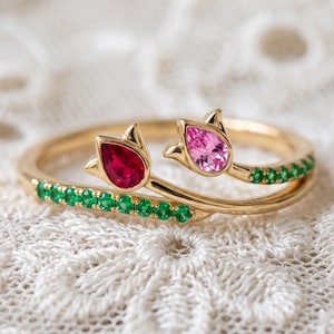 May include: A gold ring featuring two tulip-shaped settings, one holding a pear-cut red gemstone and the other a pear-cut pink gemstone. The band is accented with a row of small, round green gemstones along the shank on both sides.