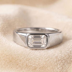 May include: A silver-toned metal ring featuring a single emerald-cut clear stone set in a bezel setting. The band is smooth and polished, with a wide, flat profile that tapers slightly towards the back, showcasing a clean and reflective surface.