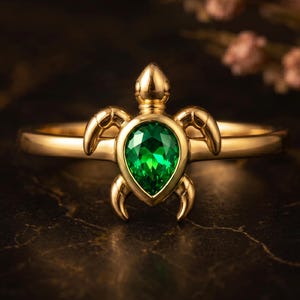 May include: A gold ring featuring a turtle-shaped centerpiece. The turtle's shell is a pear-cut, vibrant green gemstone set in a polished gold frame. The ring band is smooth and circular, showcasing a detailed head and four curved limbs.