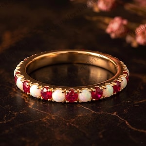 Pode incluir: Um anel de banda de ouro com um padrão alternado de rubis vermelhos redondos facetados e opalas brancas lisas, fixados numa cravação de garras partilhadas. O anel exibe uma fila contínua destas pedras preciosas em toda a sua circunferência.