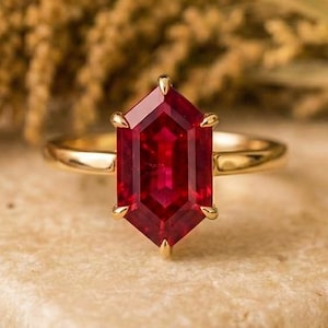 May include: A gold ring featuring a large, faceted, hexagonal ruby gemstone. The ruby is a deep red hue and is set in a gold band. The ring is displayed on a light-coloured stone surface.