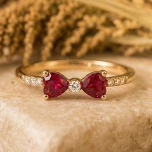 Könnte beinhalten: Ein goldener Ring mit einem Schleifendesign, das zwei herzförmige, tiefrote Edelsteine und einen zentralen Diamanten aufweist. Das Band des Rings ist mit kleinen Diamanten verziert. Der Ring liegt auf einem hellen Stein, mit einem verschwommenen Hintergrund.