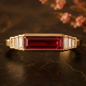 Puede incluir: Un anillo de oro con una piedra preciosa rectangular de color rojo intenso en el centro. El anillo está flanqueado por piedras preciosas blancas rectangulares más pequeñas a cada lado. El diseño se presenta sobre un fondo oscuro, resaltando los elegantes detalles del anillo.