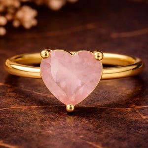 May include: A gold ring featuring a heart-shaped, light pink gemstone. The ring has a smooth, polished band and small gold accents around the heart-shaped stone. The ring is set against a dark, textured background.