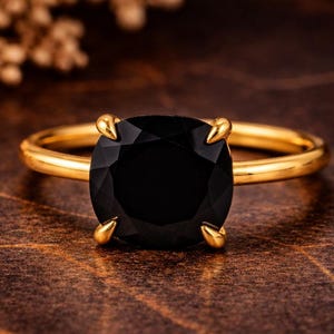 May include: A gold ring featuring a large, faceted black gemstone. The ring has a simple, elegant design with a thin gold band and four prongs holding the stone. The ring is set against a warm, brown background.