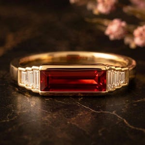 May include: A gold ring featuring a central rectangular baguette-cut red gemstone flanked by three smaller baguette-cut clear stones on each side. The band is polished gold, creating a sleek and structured design with a vibrant red focal point.