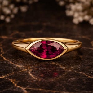 Op de afbeelding: Een gouden ring met een marquise-geslepen robijn edelsteen. De robijn heeft een dieprode kleur en is gezet in een bezel setting. De ring ligt op een donker, marmerachtig oppervlak.