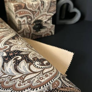 May include: A decorative tissue box and rolled fabric featuring a paisley pattern in shades of brown, beige, and black. The tissue box is square and has a matching design. A neutral-colored paper is also visible.