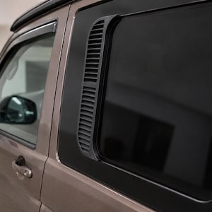 Op de afbeelding: Een metallic bronskleurig zijpaneel van een voertuig met een donker getint raam en een verticaal zwart kunststof ventilatierooster aan het frame. De buitenkant is voorzien van een bijpassende bronzen deurklink en een zwarte zijspiegel.