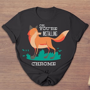 Pode incluir: T-shirt preta com uma raposa de desenho animado em tons de laranja, vermelho e branco, em pé sobre a grama verde. O texto "OH, YOU'RE INSTALLING CHROME" está impresso acima e abaixo da ilustração da raposa.