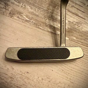May include: A silver golf putter with a black textured grip. The putter head is rectangular with rounded edges. The shaft is attached to the center of the putter head. The words "Made in USA" are printed on the shaft.