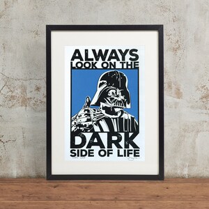 Op de afbeelding: Een zwart-witte illustratie van Darth Vader met een duim omhoog, tegen een blauwe achtergrond. De tekst "ALWAYS LOOK ON THE DARK SIDE OF LIFE" is in zwart boven Darth Vader gedrukt.