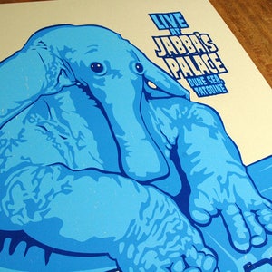 May include: A light blue poster with a stylized illustration of a creature. The poster reads "LIVE AT JABBA'S PALACE, DUNE SEA, TATOOINE" in white text. The artwork is set against a cream background.