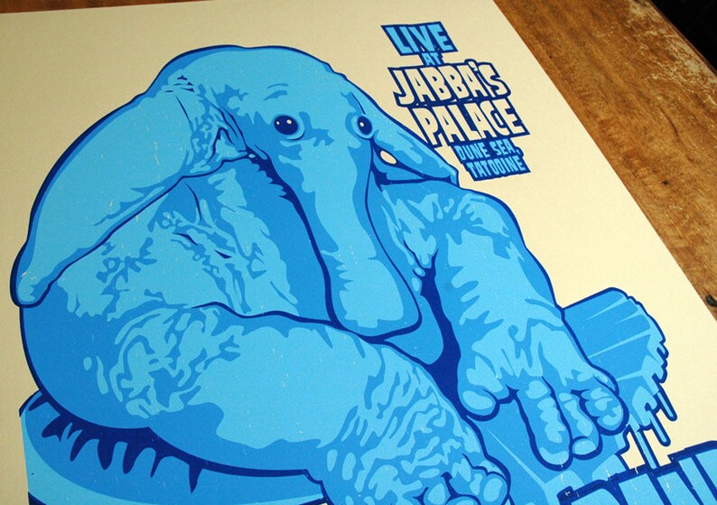 May include: A light blue poster with a stylized illustration of a creature. The poster reads "LIVE AT JABBA'S PALACE, DUNE SEA, TATOOINE" in white text. The artwork is set against a cream background.